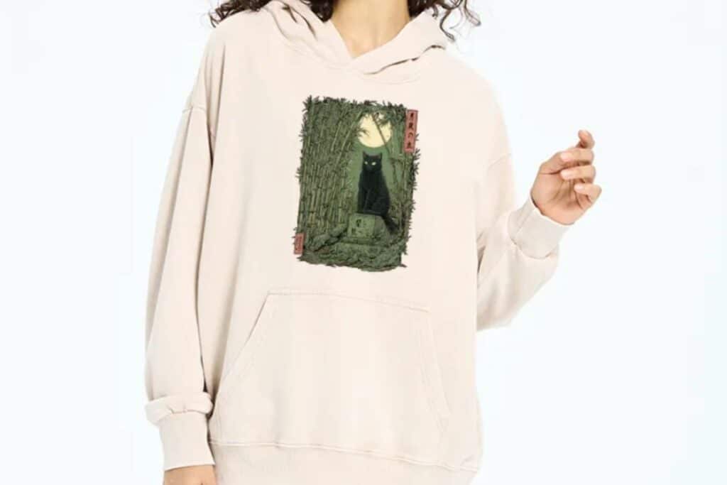 A person wearing a cream-coloured hoodie featuring an illustration of a black cat sitting on a stone path under a full moon, surrounded by tall grass and bamboo—perfect for fans of Japanese T-shirts and unique, artistic designs.