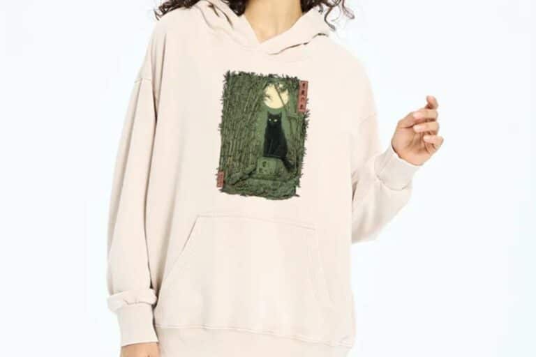 A person wearing a cream-coloured hoodie featuring an illustration of a black cat sitting on a stone path under a full moon, surrounded by tall grass and bamboo—perfect for fans of Japanese T-shirts and unique, artistic designs.