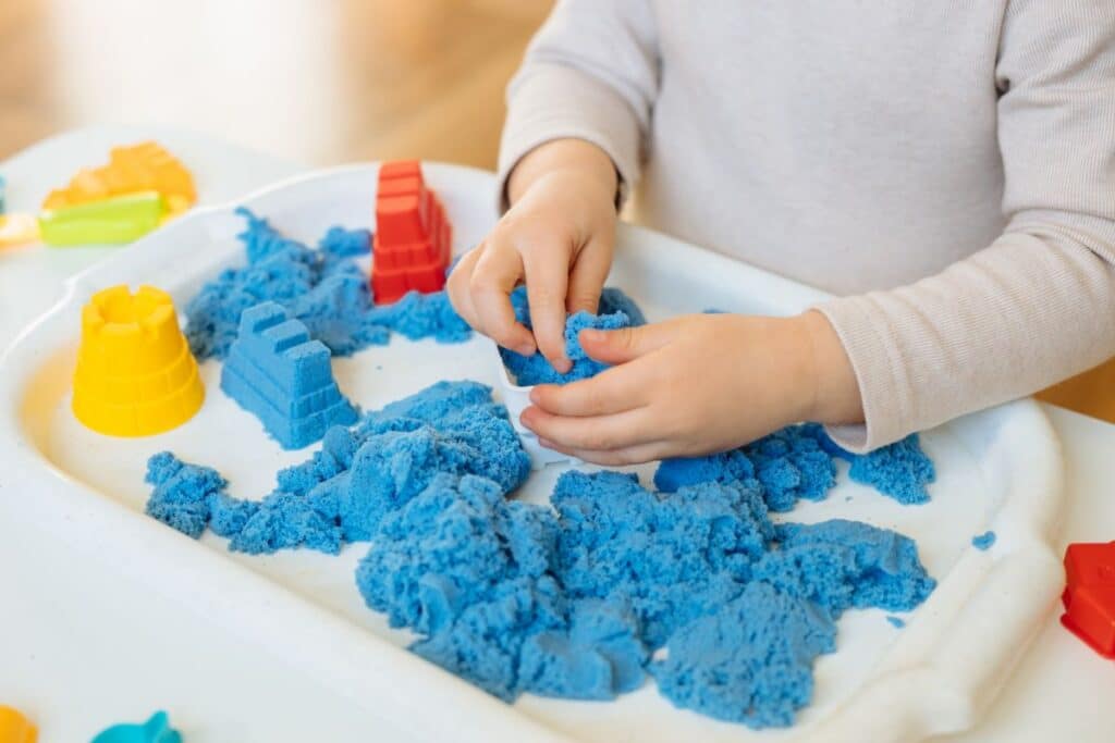 A child enjoys sensory play with blue kinetic sand on a white tray, shaping it with their hands. Colourful plastic moulds, including red, yellow, and orange shapes, are arranged around the sand.