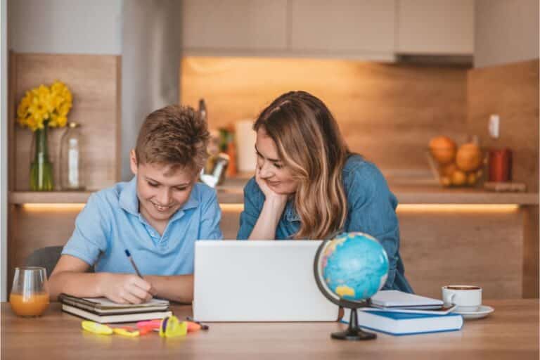 A smiling woman and boy sit at a kitchen table with a laptop, notebooks, and a small globe, practising good study habits together as they work on schoolwork or homework.
