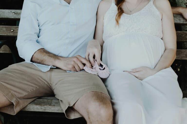 A pregnant woman in a white dress sits on a bench beside a man in shorts and a button-up shirt. Together, they hold tiny pink baby shoes on her lap, capturing the joy of becoming a parent.