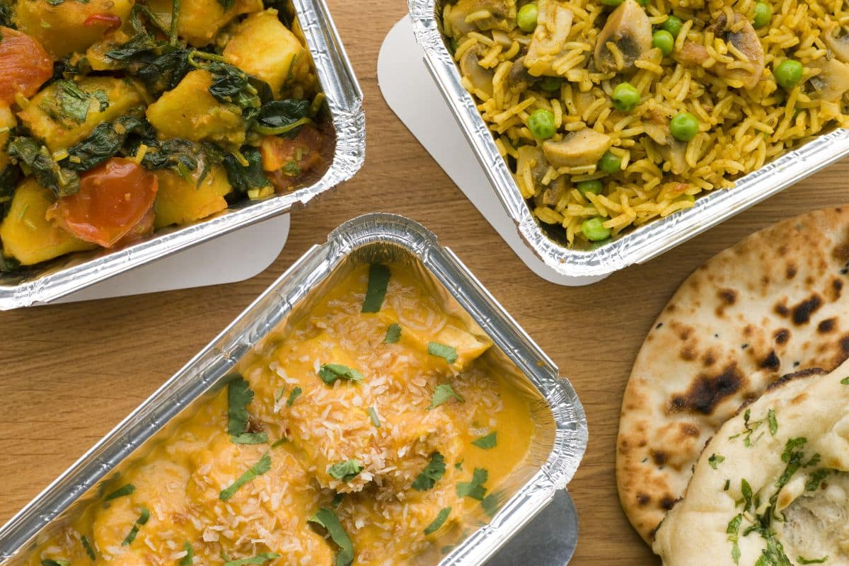 Three foil containers with Indian food—yellow curry with herbs, yellow rice with peas and mushrooms, and a vegetable dish—sit next to naan bread on a wooden surface, perfect for convenient takeaway dining.