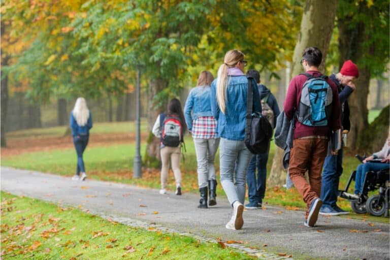 A group of people, including one person in a wheelchair, walk along a tree-lined path in a park during autumn near the uni. Many wear rucksacks and jackets, while fallen leaves cover the grass and pathway.