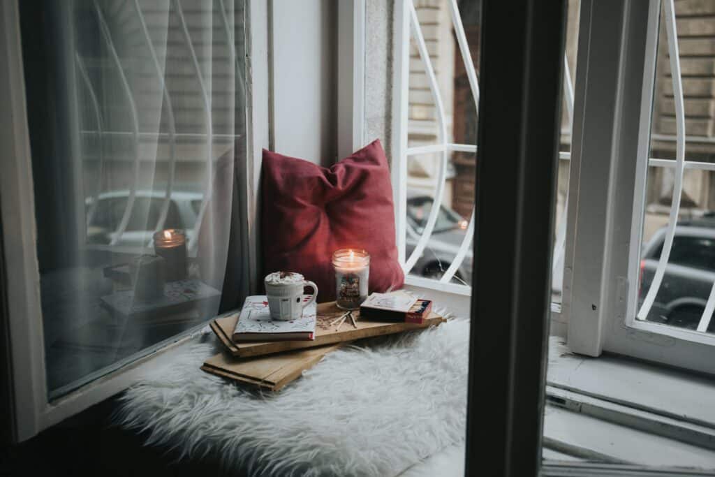 A cosy "me space at home" featuring a window seat with a red cushion, fluffy white rug, books, a mug, and a lit candle, set beside open windows overlooking buildings and parked cars.