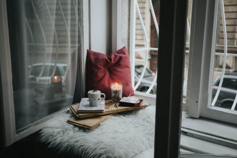 A cosy "me space at home" featuring a window seat with a red cushion, fluffy white rug, books, a mug, and a lit candle, set beside open windows overlooking buildings and parked cars.