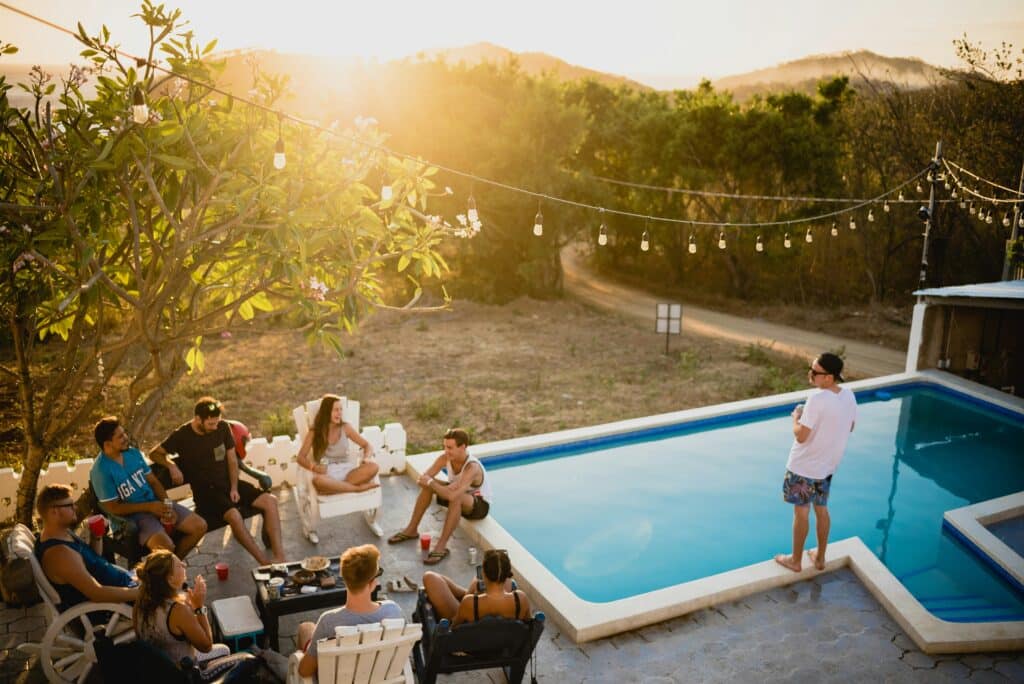 A group of people relax by the pool at sunset, sitting on chairs and benches, talking and enjoying snacks, with fairy lights overhead and trees in the background. One person stands at the pool’s edge, admiring the tranquil scene.