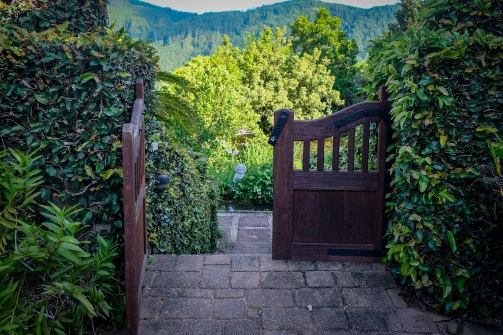 A wooden garden gate stands open on a stone path, surrounded by lush green hedges, with a view of trees and forested hills enhancing the garden’s tranquil charm.