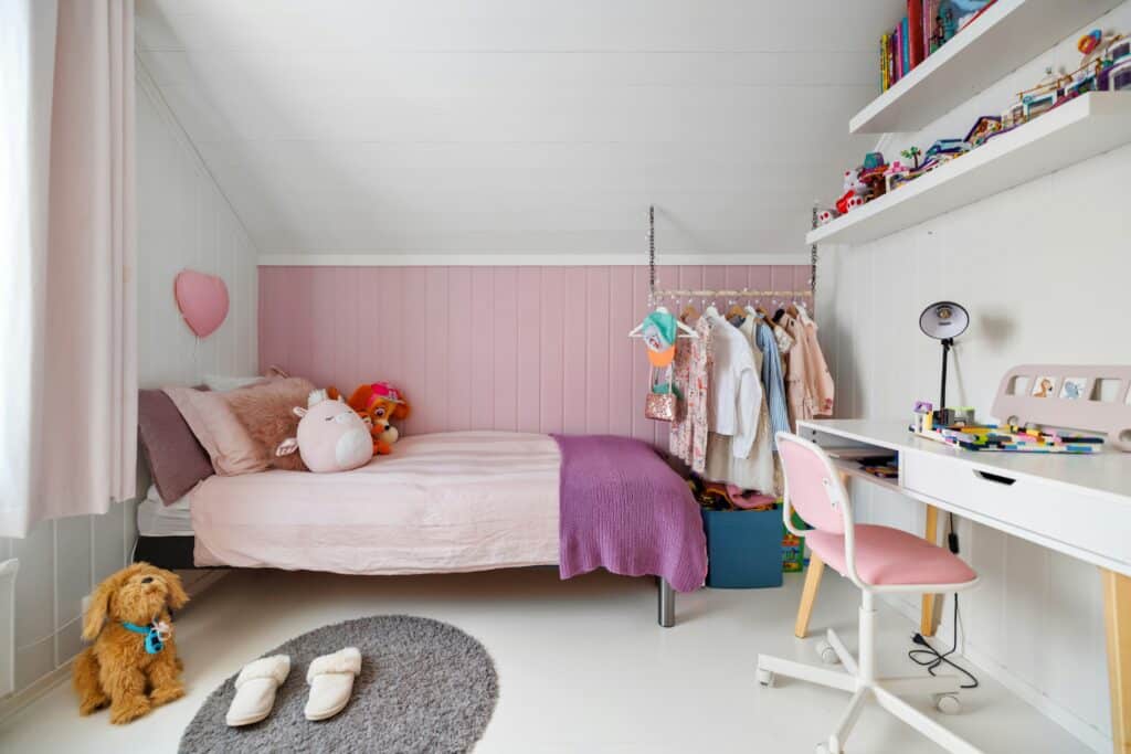 A cosy child's bedroom with a pink and white colour scheme features a bed piled with soft toys, a small desk and pink chair, a clothes rail, toy-filled shelves, slippers on a grey rug, and a plush dog on the floor.