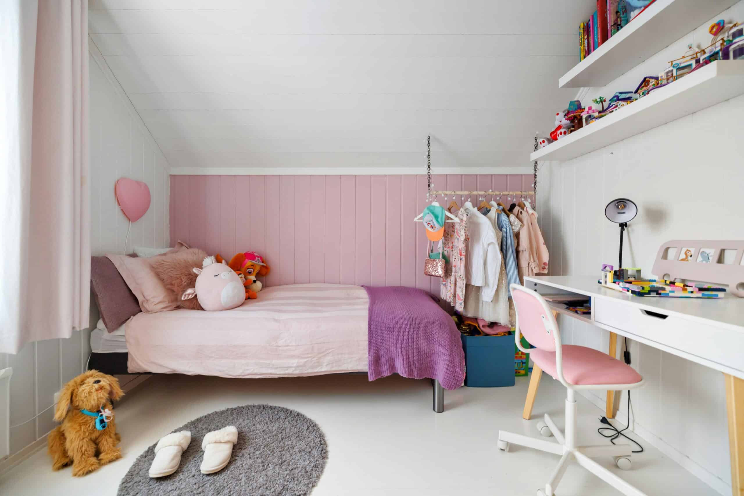 A cosy child's bedroom with a pink and white colour scheme features a bed piled with soft toys, a small desk and pink chair, a clothes rail, toy-filled shelves, slippers on a grey rug, and a plush dog on the floor.