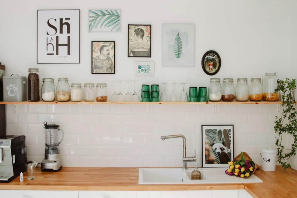 A modern kitchen with a wooden worktop, sink, blender, jars of dry goods, and green glasses on a shelf. The white kitchen wall is decorated with framed art, plants, and a bowl of colourful fruit.