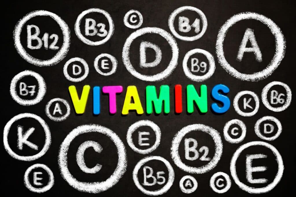 The word "VITAMINS" in colourful letters is surrounded by various vitamins, including A, B12, D, E, C, and K—all written in white chalk circles on a blackboard background.