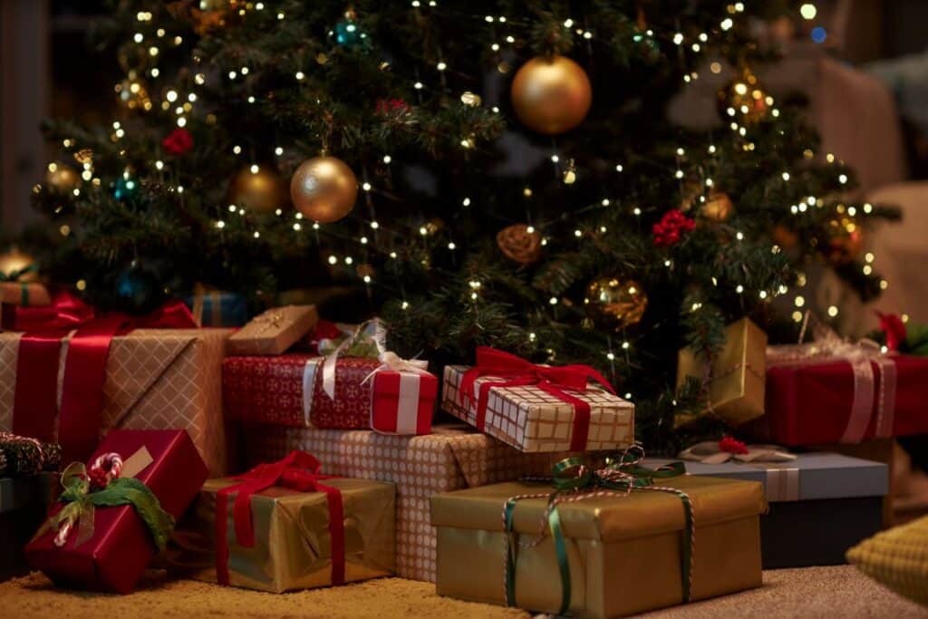 A decorated Christmas tree with gold baubles and twinkling fairy lights stands behind a pile of wrapped presents—an inspiring scene for any holiday gift guide—showcasing gifts in various sizes, patterns, and colours, all tied with ribbons and bows.