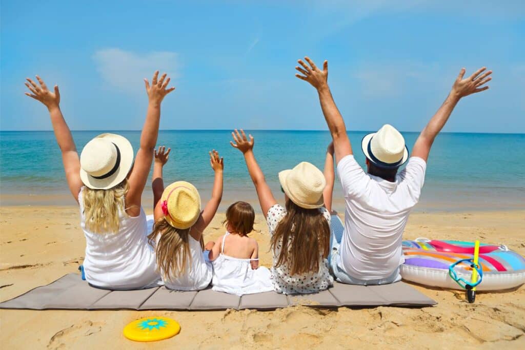 A family of five, all wearing hats, sit on a mat facing the sea. They raise their arms in joy on a sandy beach under a sunny sky, with beach toys and an eSIM-ready mobile beside them.