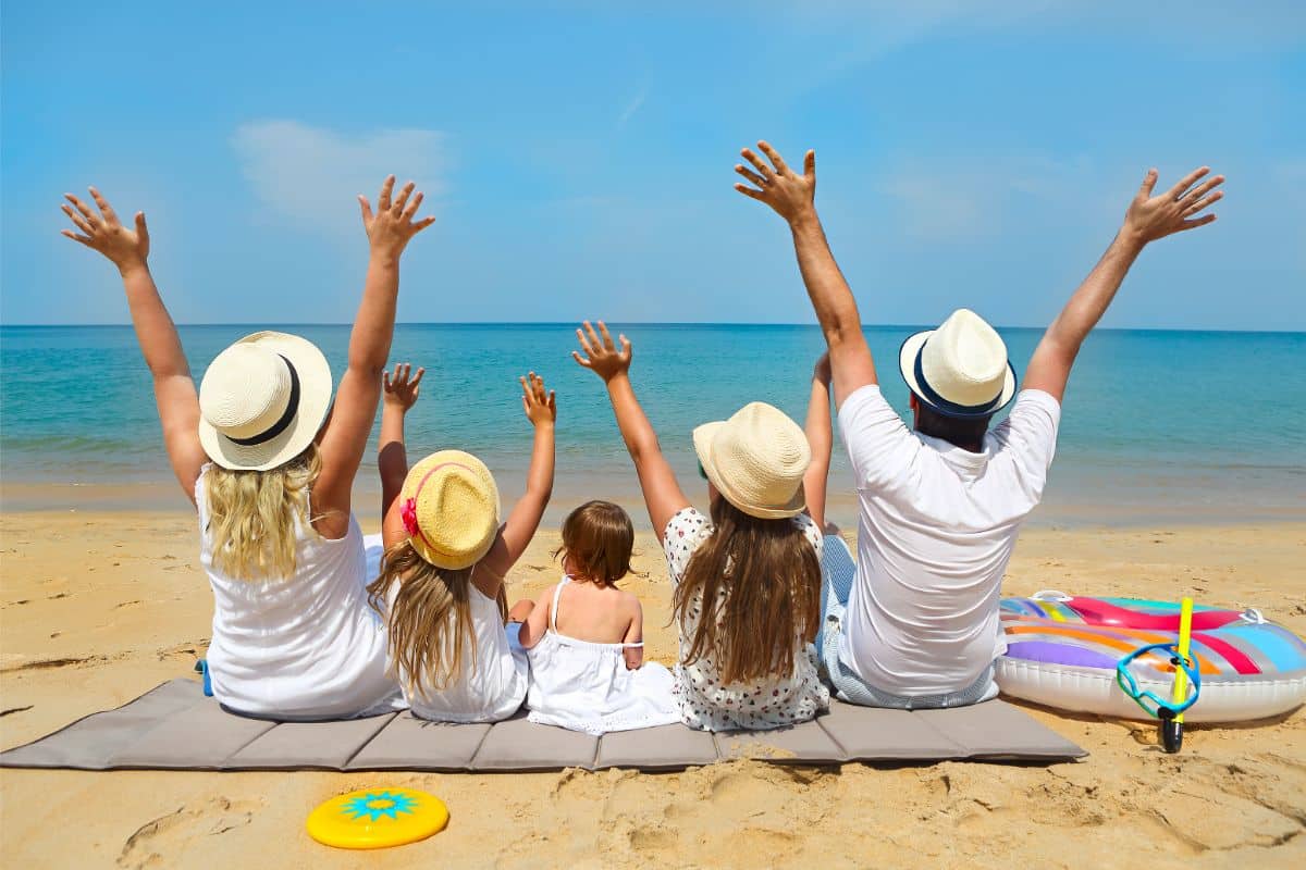 A family of five, all wearing hats, sit on a mat facing the sea. They raise their arms in joy on a sandy beach under a sunny sky, with beach toys and an eSIM-ready mobile beside them.