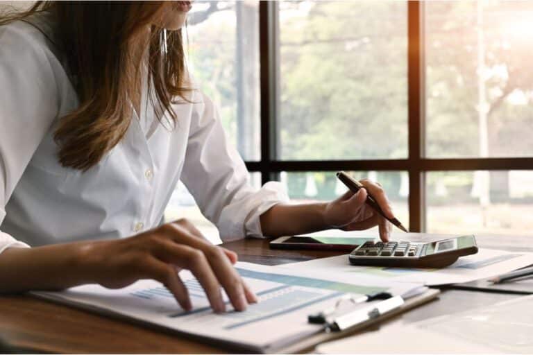 A woman sits at a desk using a calculator and reviewing car finance claims alongside financial documents with charts and graphs, in front of large windows letting in natural light.
