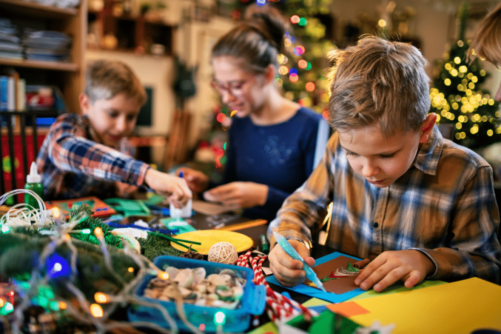 Three children sit at a table, focused on making holiday crafts with paper and felt-tip pens. Christmas lights and a decorated tree in the background highlight the joy of family Christmas traditions in a festive atmosphere.