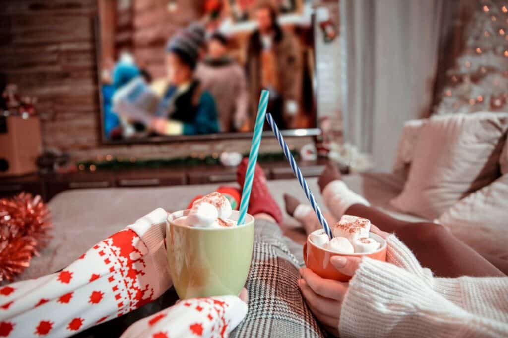 Two people wearing cosy winter clothes hold mugs of hot chocolate with marshmallows and straws, enjoying a holiday film on TV in a festive sitting room decorated with Christmas lights and a tree—a perfect moment of family Christmas traditions.