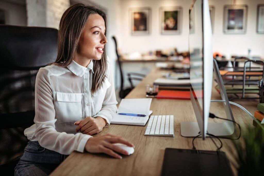 A woman in a white blouse sits at a desk, using a computer mouse and looking at a desktop monitor, researching traffic strategy. A notebook, pen, and keyboard rest on the desk in this modern office setting.
