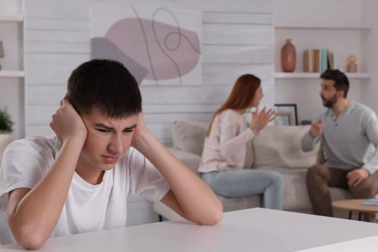 A teenage boy sits at a table covering his ears, looking upset, whilst a man and woman argue in the background on a sofa in a living room.