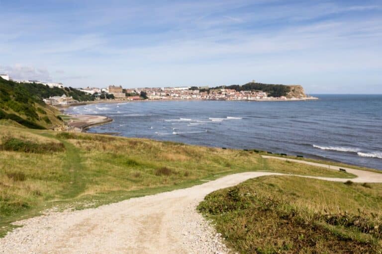 A winding path leads through grassy Yorkshire hills towards a coastal town by the sea, perfect for budget-friendly family days out, with waves gently breaking on the shore and buildings clustered near a distant headland under a blue sky.