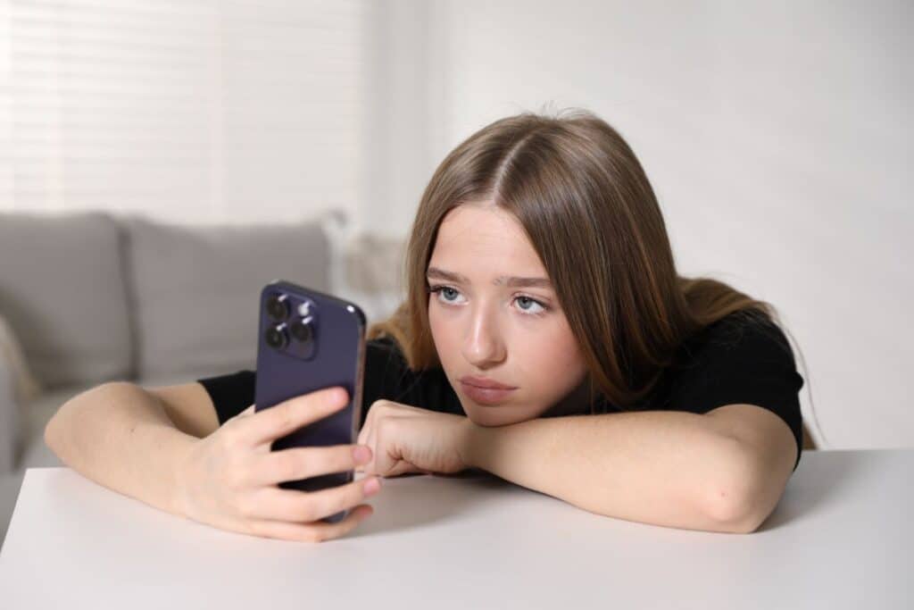 A young teen with long brown hair rests her head on her arm, looking at a mobile phone with a sad or bored expression whilst sitting at a white table in a bright room—perhaps searching for indoor activities on rainy days.