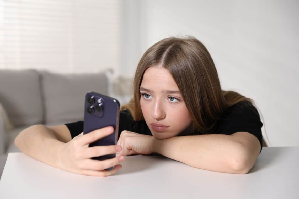 A young teen with long brown hair rests her head on her arm, looking at a mobile phone with a sad or bored expression whilst sitting at a white table in a bright room—perhaps searching for indoor activities on rainy days.