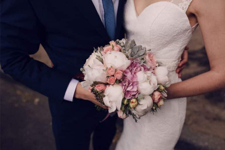 A bride in a white lace dress holds a bouquet of pink and white flowers, standing close to a groom in a dark suit and blue tie. Their embrace captures meaningful moments that reflect their unique wedding style. Faces are out of shot.