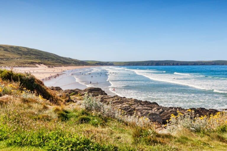 A scenic view of a sandy beach in Devon with people enjoying a family holiday by the water and shore, gentle waves rolling in, grassy dunes and rocky outcrops in the foreground, and green hills in the distance under a clear blue sky.