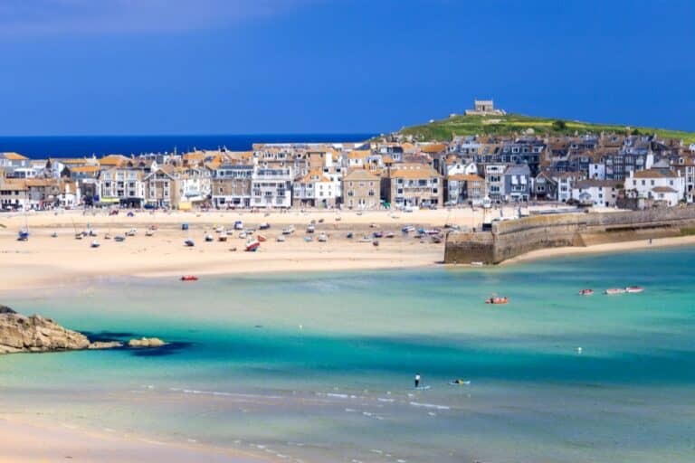 A coastal town in Cornwall with sandy beaches, turquoise water, and small boats. Rows of white and stone houses line the shore, perfect for family holidays, with a grassy hill and blue sky in the background.