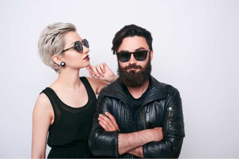 A woman with short blonde hair and a man with a beard, both in bold streetwear—black sunglasses and black outfits—stand together against a plain white background. The woman looks at the man, touching his shoulder.