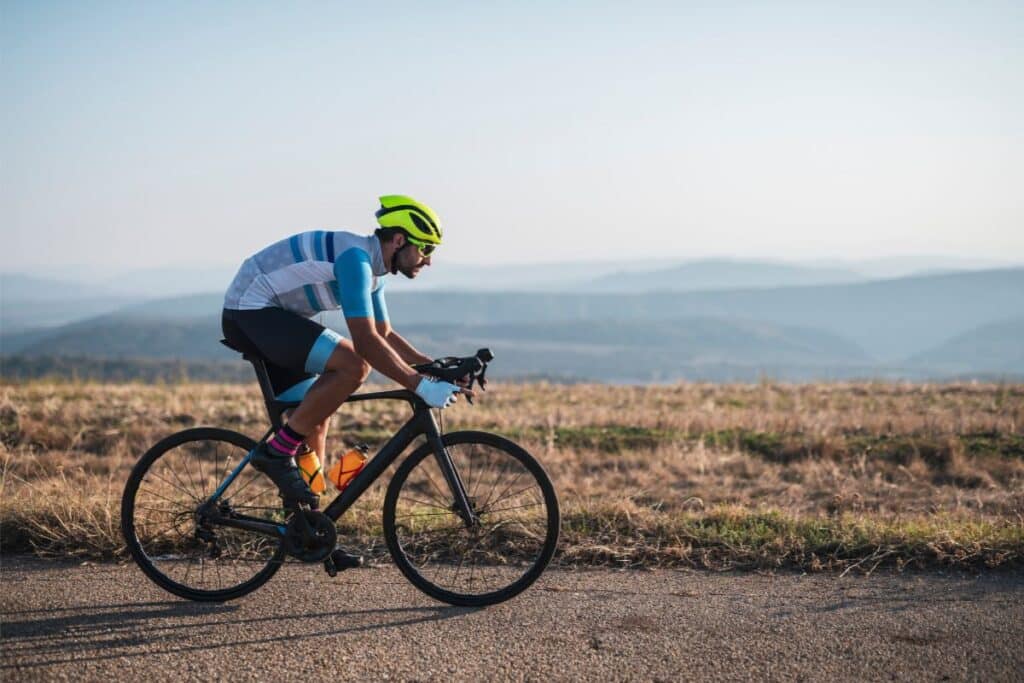 A cyclist wearing a bright yellow helmet and blue cycling apparel rides a road bike on a tarmac path, with dry grass and distant mountains visible under a clear sky—perfect inspiration for bike gifts.