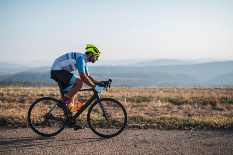 A cyclist wearing a bright yellow helmet and blue cycling apparel rides a road bike on a tarmac path, with dry grass and distant mountains visible under a clear sky—perfect inspiration for bike gifts.