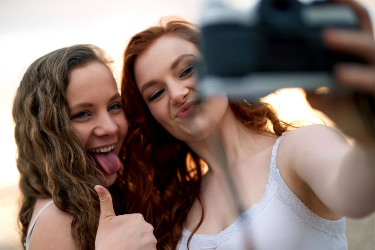 Two young women taking a selfie together, smiling and pulling playful faces—one sticks out her tongue and gives a thumbs up, while the other does a duck face—a perfect moment for any teen bucket list. The background is softly blurred.