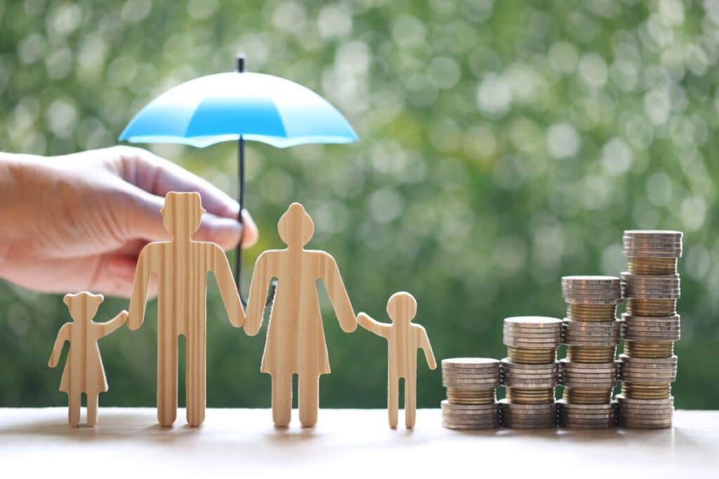 A hand holds a blue umbrella over wooden cut-outs of a family of four, beside stacks of coins, symbolising how to protect finances and ensure security for family finances.