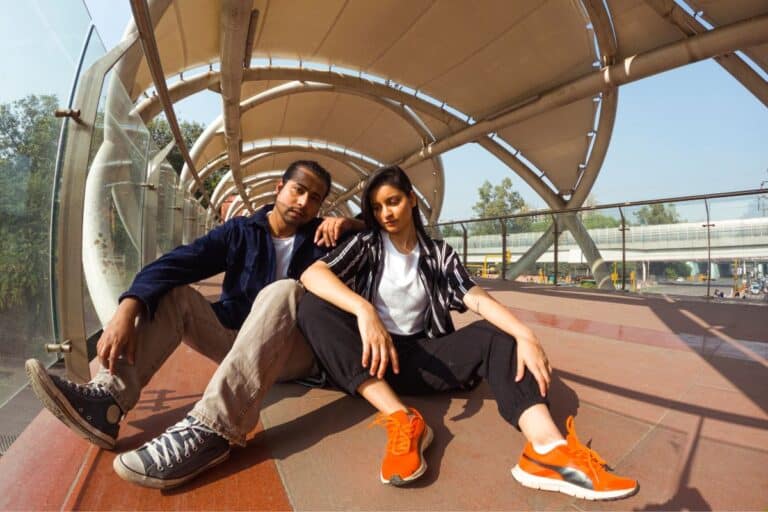 Two young adults sit on the ground in a modern, covered walkway, posing confidently. Both wear casual clothes and trainers, with sunlight streaming through the glass structure behind them.