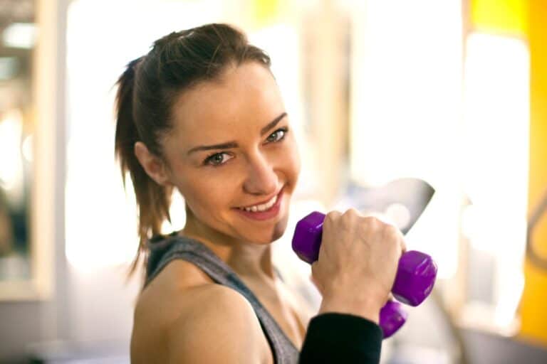 A woman in gym clothing smiles at the camera whilst holding a purple dumbbell, standing in a brightly lit gym or fitness studio.
