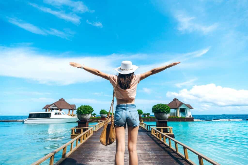 A woman in a hat stands on a pier with arms outstretched, facing the sea, embracing a unique travel experience. Overwater bungalows, a boat, and a bright blue sky create the perfect backdrop for this exclusive holiday retreat.