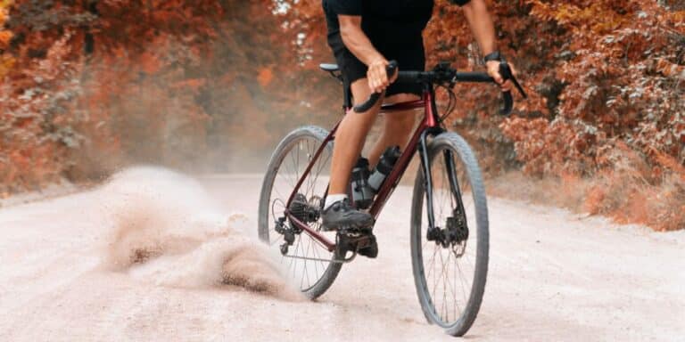 A cyclist, dressed in black and embracing comfortable riding essentials, rides a red bike on a dusty road, causing dirt to spray up behind the rear wheel. Autumn-coloured trees line both sides of the road, capturing the true spirit of gravel cycling.