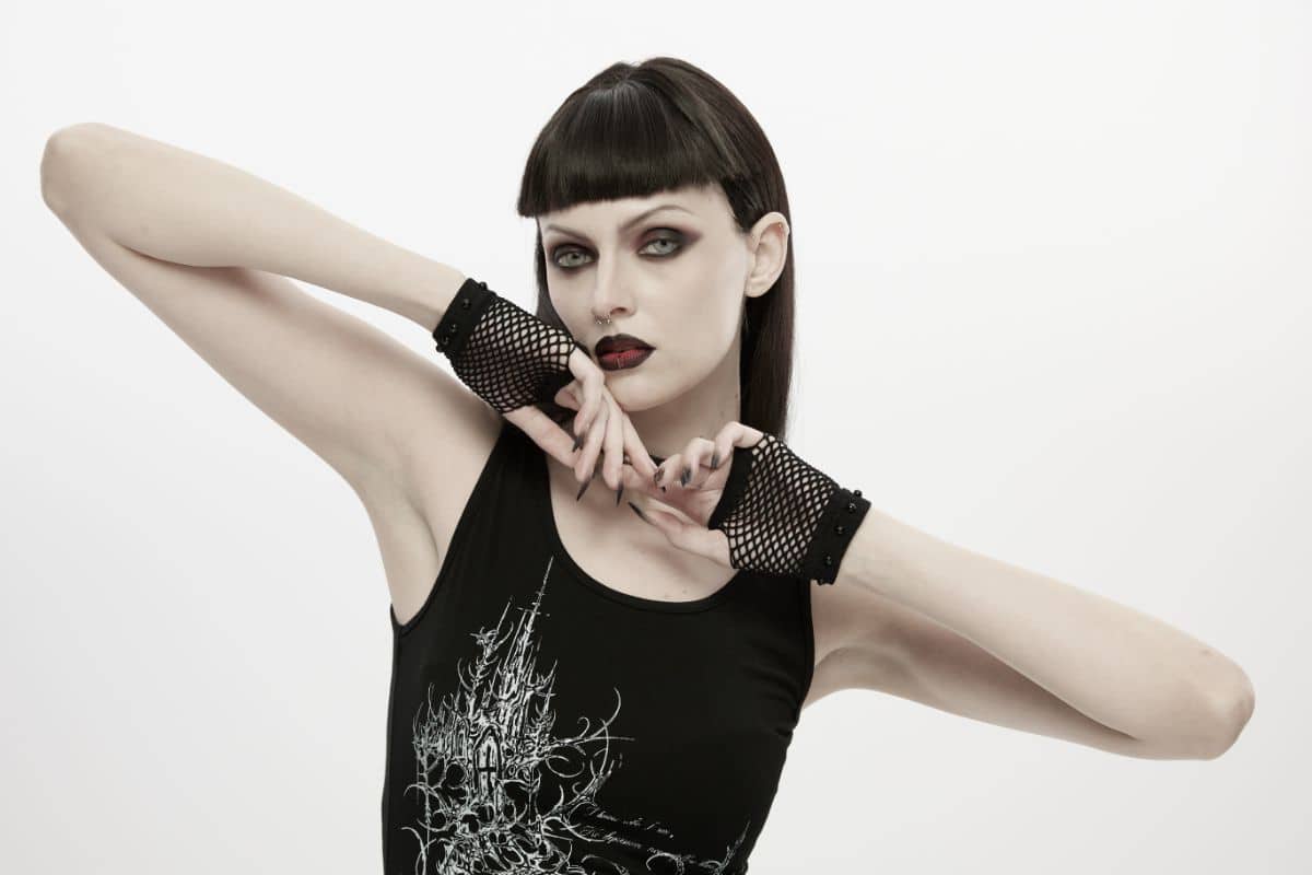 A woman with dark make-up and black lipstick poses with her arms raised, showcasing her unique style. She wears a black sleeveless top with a white pattern and black fishnet fingerless gloves, set against a plain white background—a bold nod to alternative fashion.
