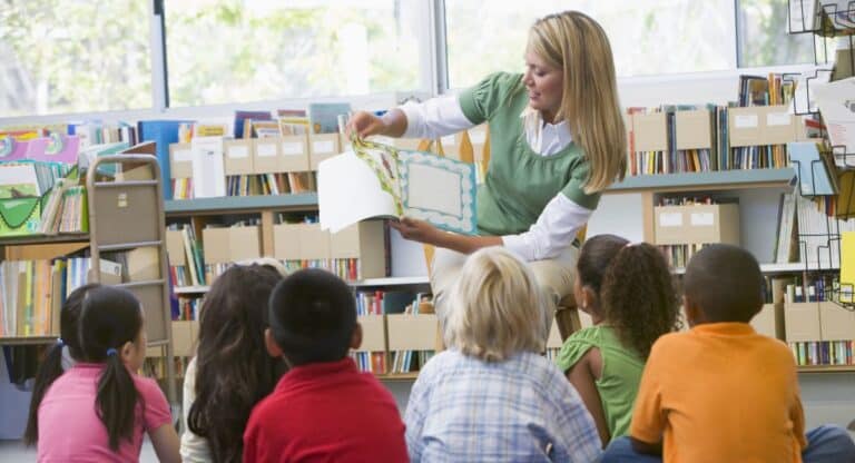 A woman, dressed in stylish teacher attire, sits in front of a group of young children, holding up a picture book and reading aloud in a library or classroom filled with bookshelves.