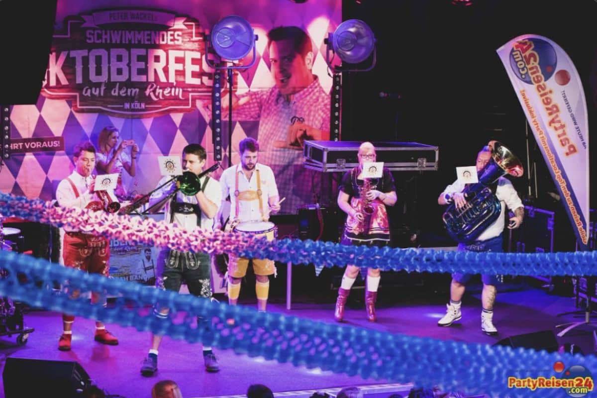 A German band in traditional lederhosen and dirndls plays festive music on a stage in front of a large Oktoberfest banner.